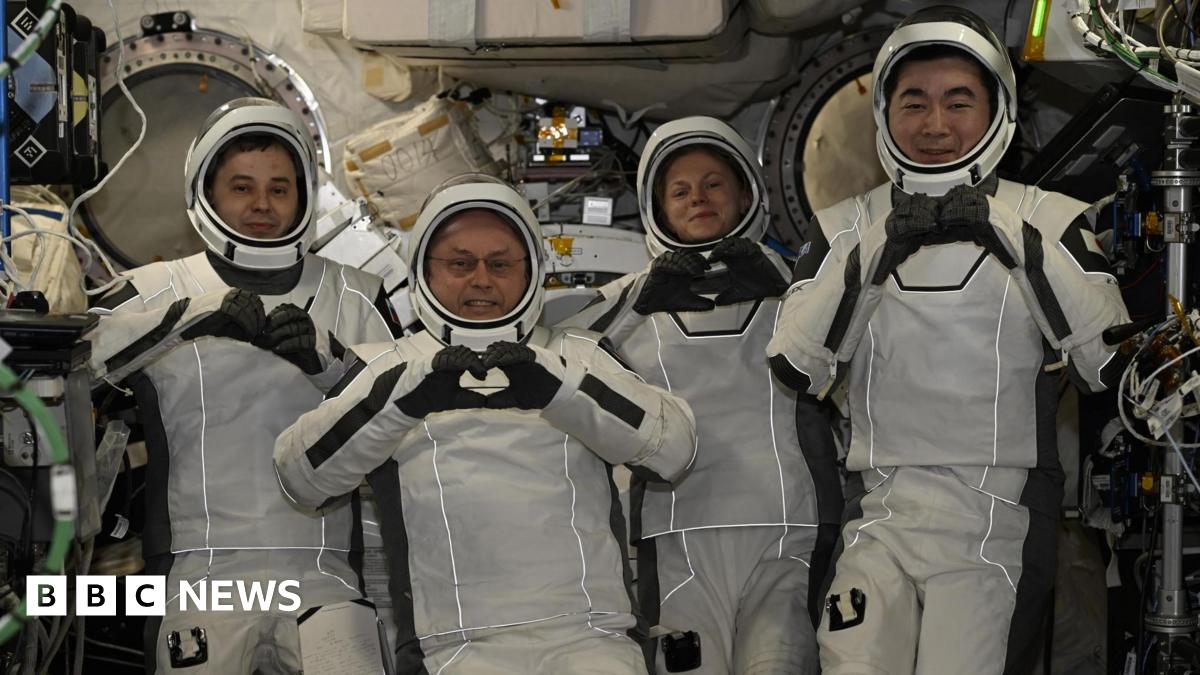 Four astronauts wearing white spacesuits and helmets pose for cameras smiling and making heart shapes with their hands and fingers. They are surrounded by wires and other equipment inside the International Space Station.