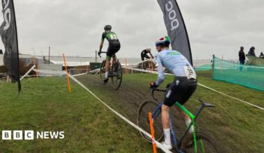 Two bike-riders taking part in the 2026 Lloyds National Cyclo-cross Championships are racing up a muddy hill between two tape fences.