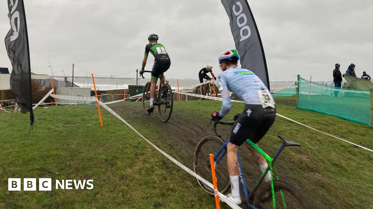 Two bike-riders taking part in the 2026 Lloyds National Cyclo-cross Championships are racing up a muddy hill between two tape fences.