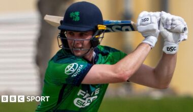 Lorcan Tucker, in dark blue helmet and mainly green cricket shirt, watches the ball after playing a shot, with the bat over his left shoulder as he looks to his right
