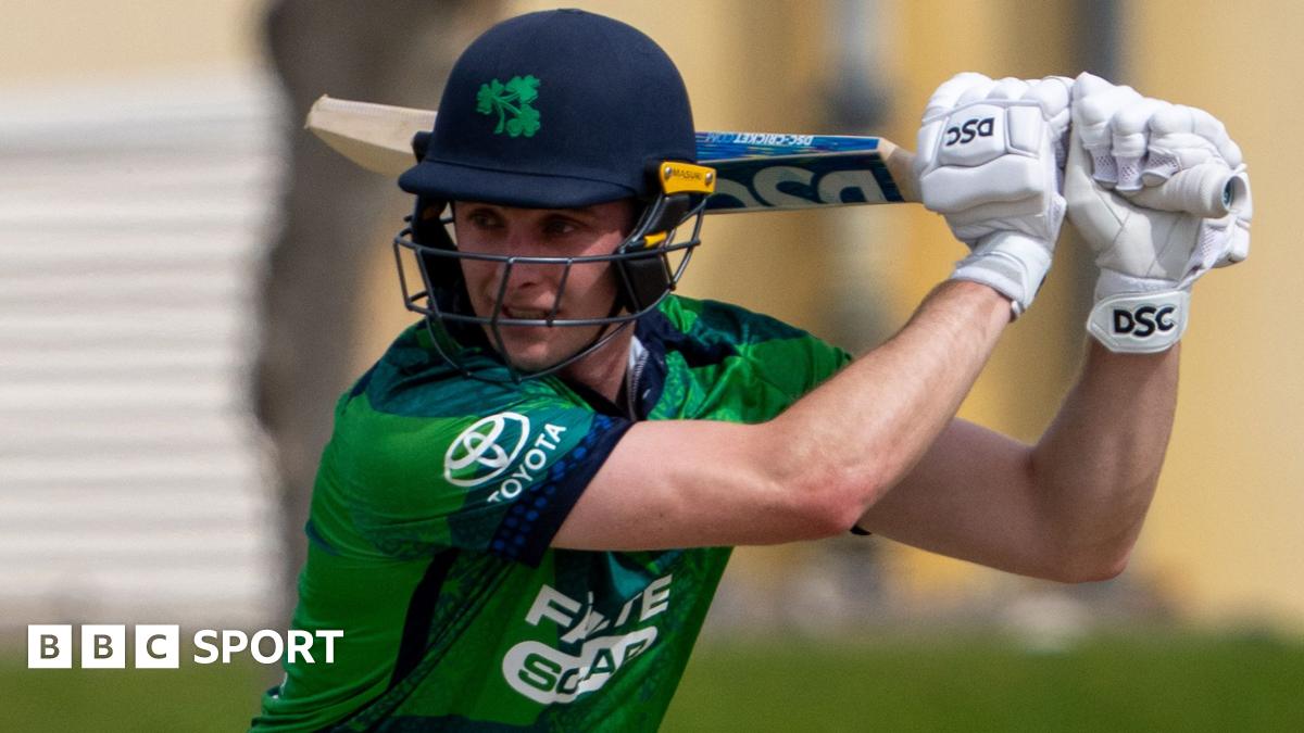 Lorcan Tucker, in dark blue helmet and mainly green cricket shirt, watches the ball after playing a shot, with the bat over his left shoulder as he looks to his right