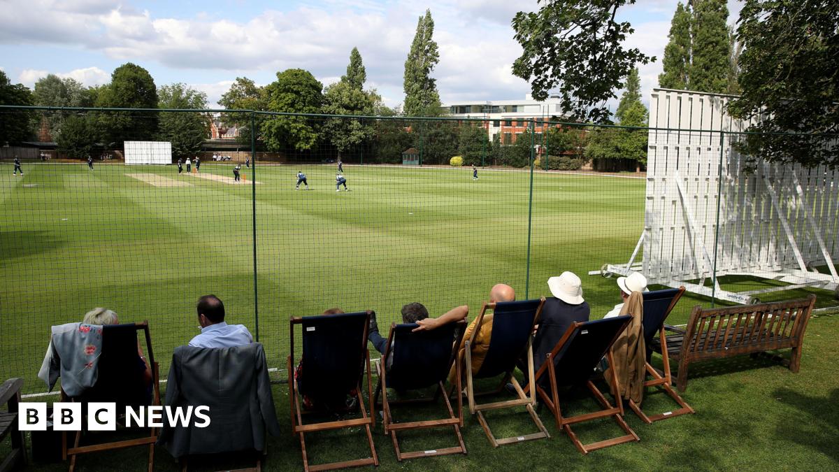 Guests sitting in deckchairs watch a cricket match from behind a black net. A white sight screen is to the right and players can be seen gathered around the batter on a brown cricket strip surrounded by green grass