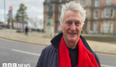 Mike Lloyd with grey hair in a blue jacket, wearing a red scarf, smiles at the camera. He is standing outside in front of a large building, which is on the other side of the road he is standing next to.