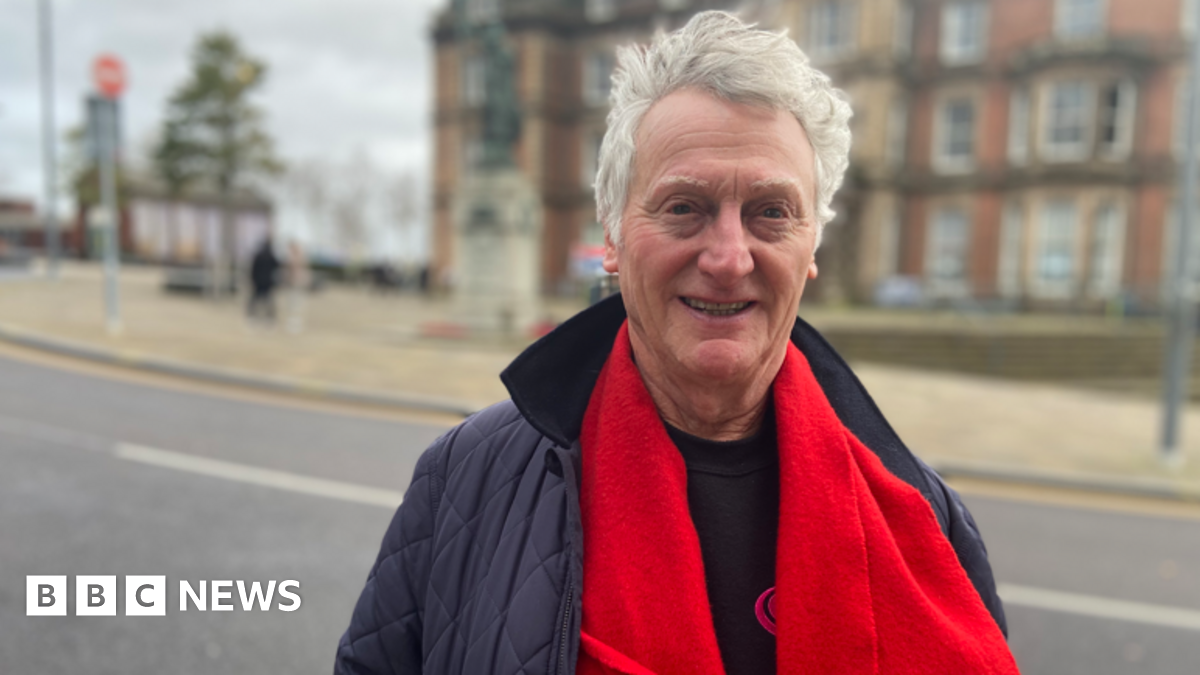 Mike Lloyd with grey hair in a blue jacket, wearing a red scarf, smiles at the camera. He is standing outside in front of a large building, which is on the other side of the road he is standing next to.