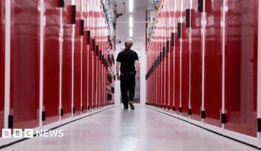 Millie Bobby Brown and David Harbour in character as Eleven and Jim Hopper. Eleven wears red shorts over grey leggings and a grey jumper. Jim Hopper wears a black leather jacket, a black beanie hat and dark trousers.They are speaking to director Frank Darabont on the set of Stranger Things. There is a vintage advertisement posted in the background with debrief on the ground.
