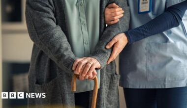 A care worker is holding a patients arm. The patient has both there hands on top off a wooden stick. The care worker is dressed in blue. The patient is wearing a green shirt with grey cardigan.