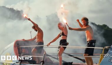 Three rowers stand on top of boat in swimwear holding flaming flares aloft in celebration
