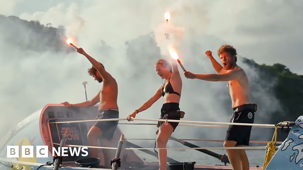 Three rowers stand on top of boat in swimwear holding flaming flares aloft in celebration