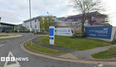 The front entrance of a hospital with large white buildings and a small roundabout in front of a glass entrance building. There is a blue and white sign at the front that reads "The Robert Jones and Agnes Hunt Orthopaedic Hospital"