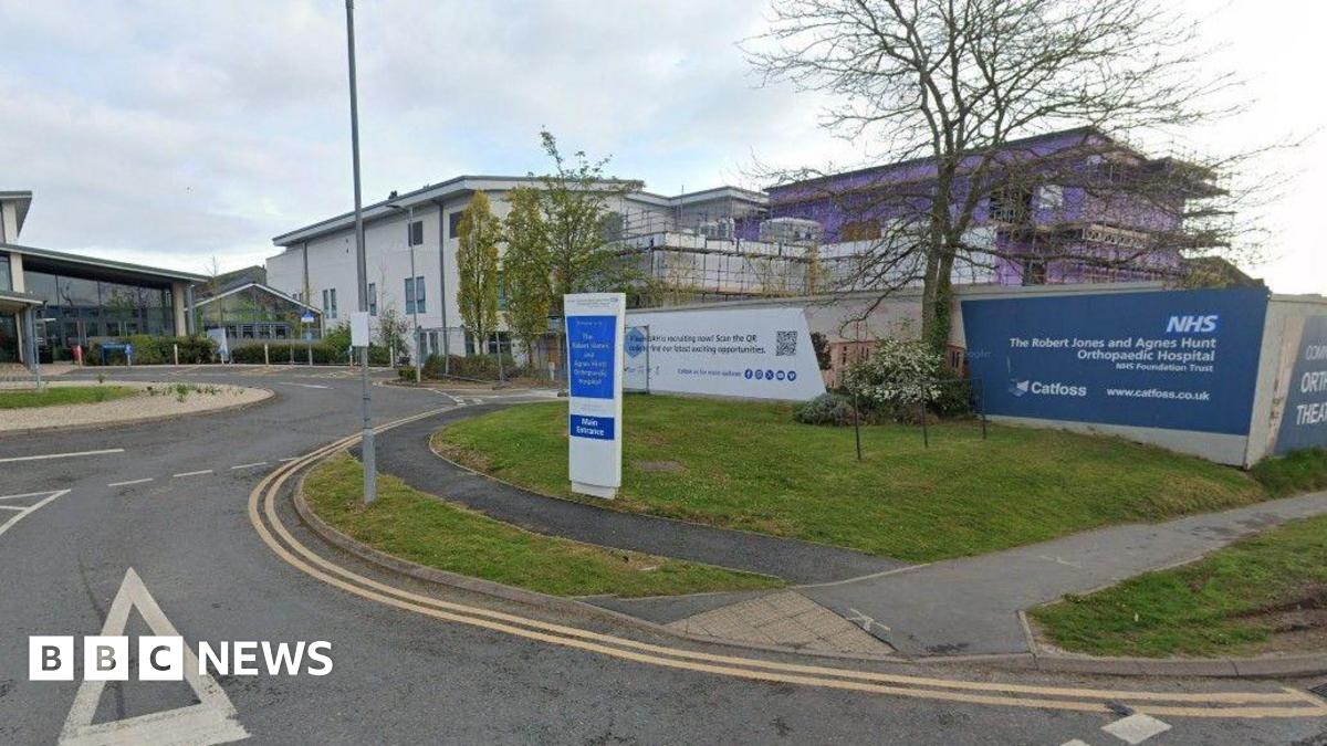 The front entrance of a hospital with large white buildings and a small roundabout in front of a glass entrance building. There is a blue and white sign at the front that reads "The Robert Jones and Agnes Hunt Orthopaedic Hospital"