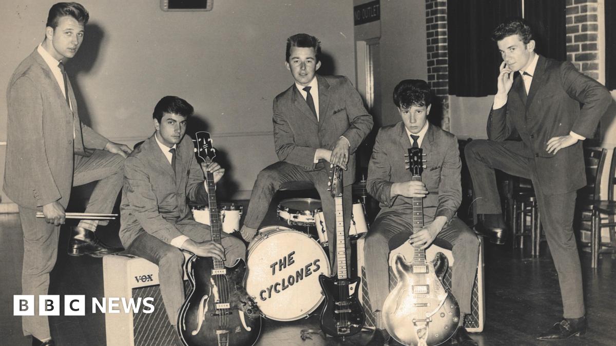 Five young men photograhped in the 1950s wearing suits typical of that period. Two are holding guitars, one is standing over a drumkit with "The Cyclones" written on the front.
