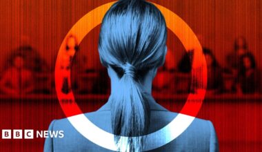 Back view of a woman with long hair in a ponytail, dressed in a dark suit, standing before a blurred red-toned background resembling a courtroom or formal panel. She is framed within a circular graphic element, partially red and white, creating a dramatic and symbolic visual effect. The overall atmosphere suggests seriousness and tension, possibly related to legal proceedings or justice.