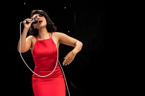 Reuters Raye sings on stage wearing a red dress with a microphone in her hand, with the lead over her shoulder 