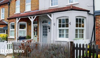 A row of terraced houses with brick and painted facades, white-framed windows, small front gardens, and low wooden fences along a residential street.