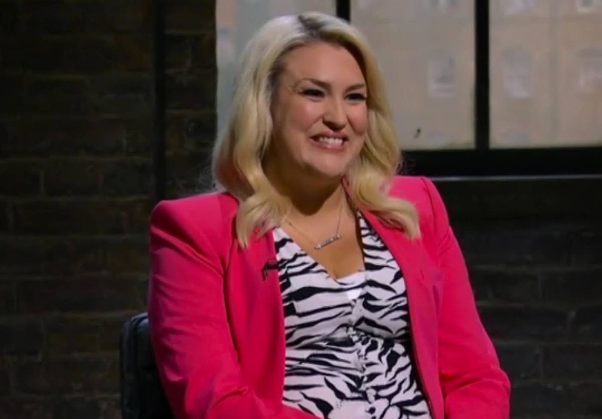 An individual with blonde hair wearing a pink blazer over a zebra-print top, seated and smiling against a brick wall background with a window.
