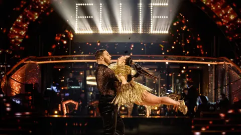 Ellie is wearing a gold sparkly dress and trainers. Her feet are off the ground as she is spun around by Vito, who is wearing dark trousers and a dark top. Ellie's name is in lights in the background, with the Strictly band sitting below
