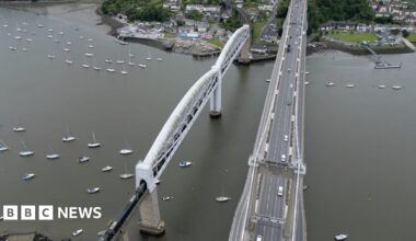 Bird's eye view of the Tamar bridge with toll booths at one end and several vehicles travelling along it.
