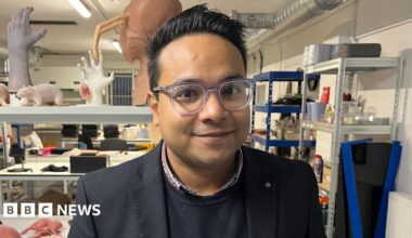 Nazmus Tareque from Anthrotek. He is standing inside an industrial unit where there are rows and tables with prosthetic items including silicon rats, body parts and organs. He is looking directly at the camera and smiling and is wearing a red and blue shirt with a blue jumper and dark blazer over the top.