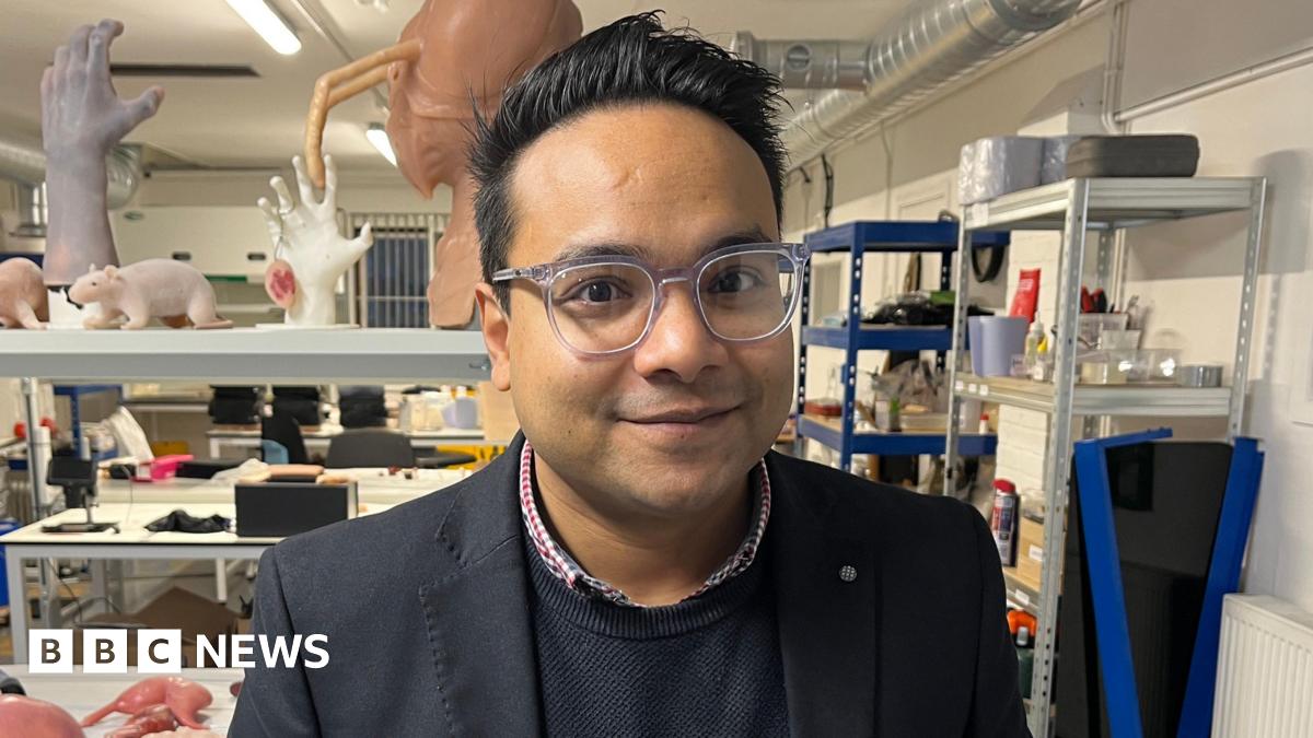 Nazmus Tareque from Anthrotek. He is standing inside an industrial unit where there are rows and tables with prosthetic items including silicon rats, body parts and organs. He is looking directly at the camera and smiling and is wearing a red and blue shirt with a blue jumper and dark blazer over the top.