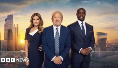 Lord Alan Sugar wears a blue suit and blue tie. He is flanked on his left by business executive Karen Brady, dressed in a blue dress, and on his right by former winner Tim Campbell, also wearing a blue suit and blue tie. They are stood in front of a backdrop of the city of London.
