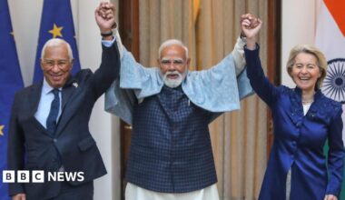 Three leaders hold hands and raise their arms, laughing during a friendly moment. The man in the centre, Indian Prime Minister Narendra Modi, with a white beard and glasses, wears a light shawl over a dark outfit. On either side, European Council President António Costa in a dark suit and European Commission President Ursula von der Leyen in a dark blue long jacket