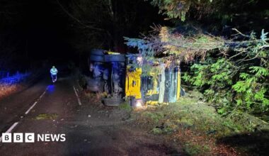 A gritter lorry on its side on a roadside in the early hours. It is dark and there is lighting on it.