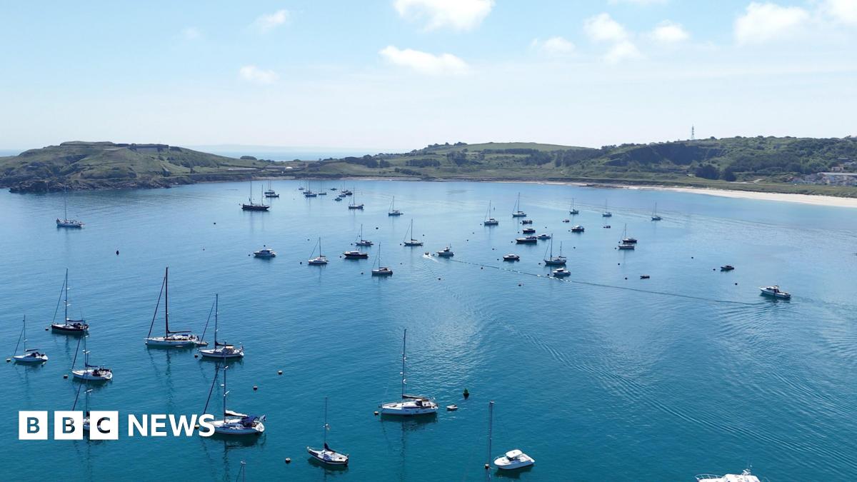 Image shows yachts on calm blue waters with golden sand and green coastline of the island of Alderney.