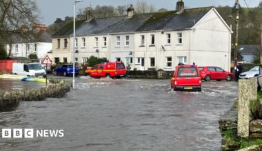 Red vehicles parked at the top of a hill as flood water runs down the road. One of the vehicles, to the right, is moving through the flood water.