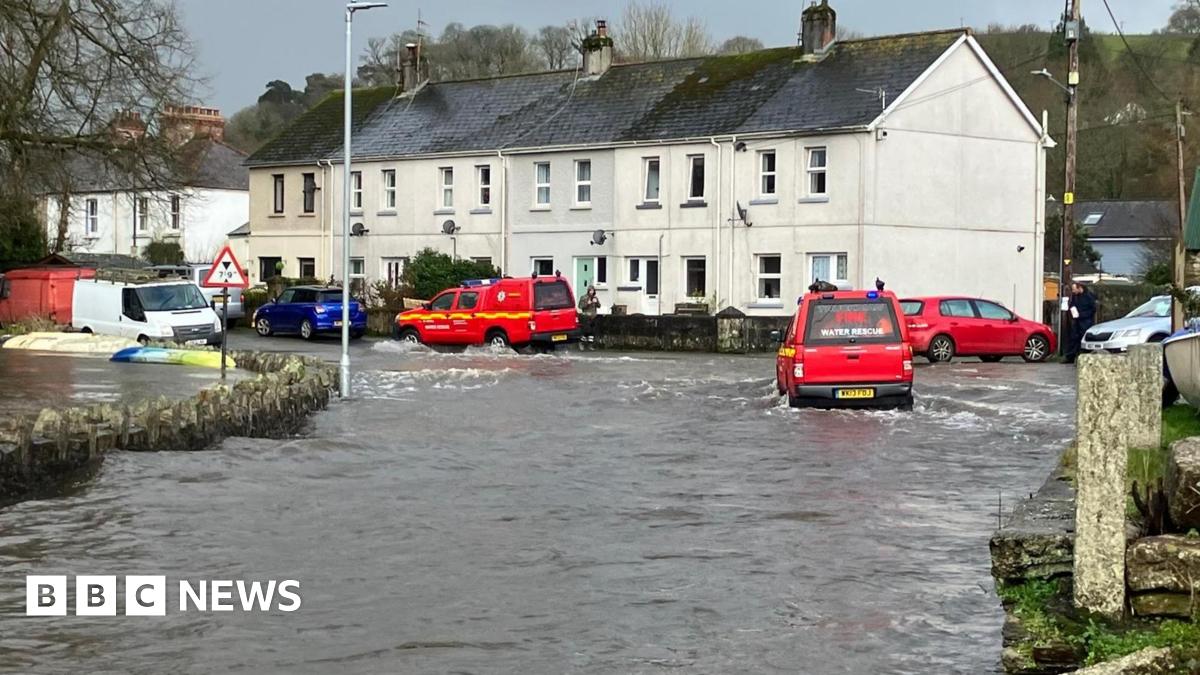 Red vehicles parked at the top of a hill as flood water runs down the road. One of the vehicles, to the right, is moving through the flood water.