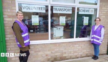 Two women in purple high-vis jackets. They are stood beside a window which has signs up that read 'Volunteer Transport Scheme'. They are looking at teh camera and smiling.
