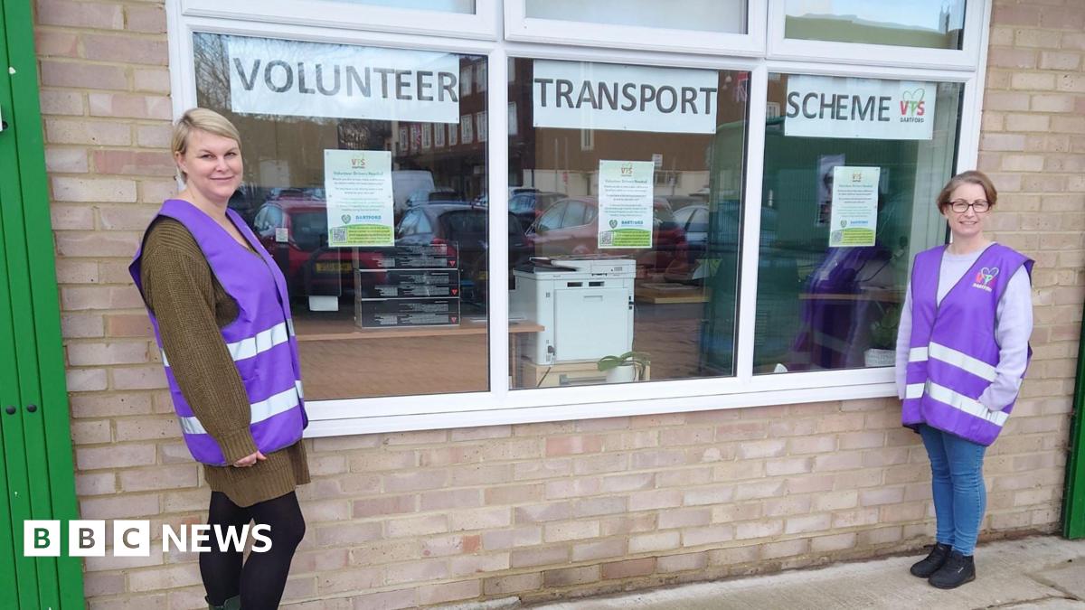Two women in purple high-vis jackets. They are stood beside a window which has signs up that read 'Volunteer Transport Scheme'. They are looking at teh camera and smiling.