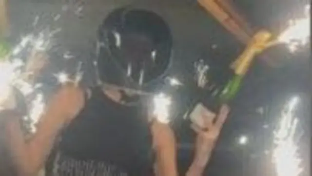 A person in  a crash helmet holding a bottle of champagne with a sparkler attached