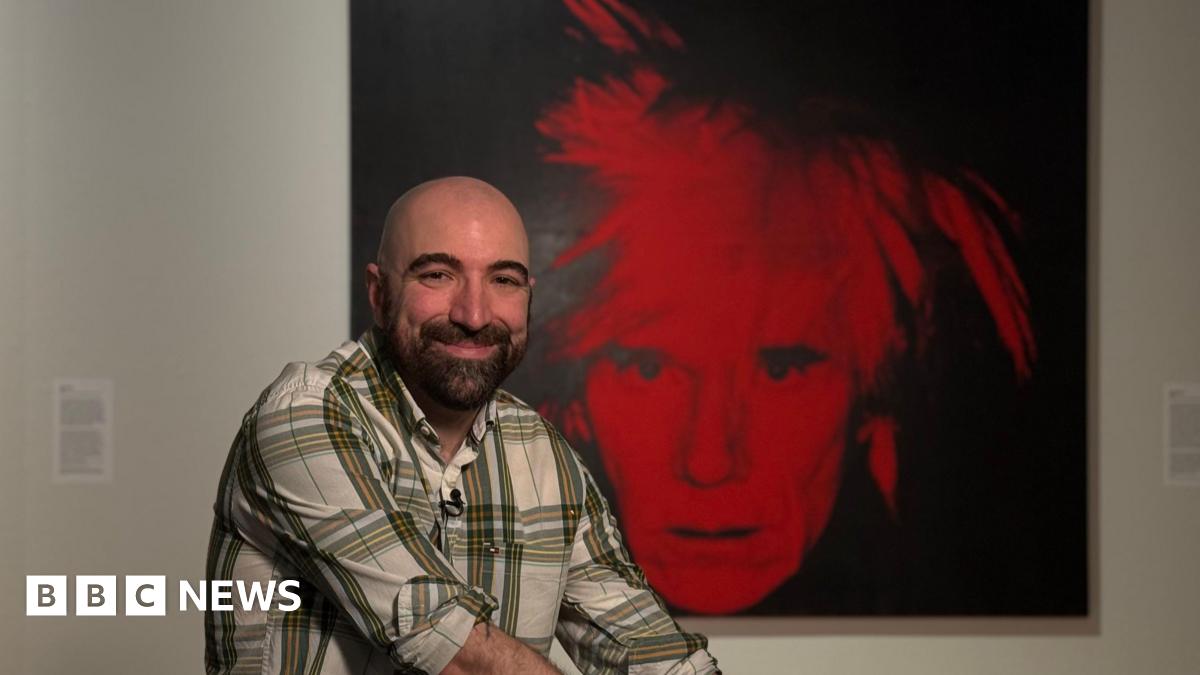 A man smiling in a gallery in front of a black and white self portrait by Andy Warhol