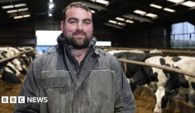 Gethin Hughes, dairy farmer stands in his cattle shed wearing blue overalls