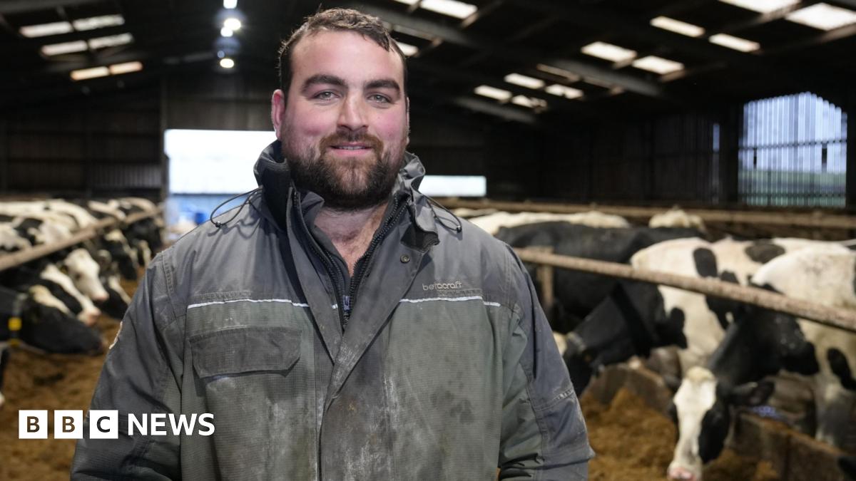 Gethin Hughes, dairy farmer stands in his cattle shed wearing blue overalls