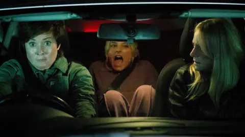 ITV Coronation Street characters Becky, Betsy and Lisa in a car at night, with Becky driving and looking startled, and Betsy in the back seat screaming