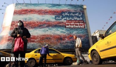 A woman is seen carrying a shopping bag and a man is seen getting into a yellow taxi cab in the foreground of the photo, while in the background there is a huge anti-US billboard which reads 'If you sow the wind, you will reap the whirlwind'