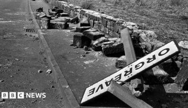 A black and white photo from 1984 of the Orgreave town road sign on the floor in a pile of rubble. A wall next to a road is torn down and the sign in lying on stones from the broken wall.