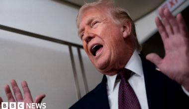 US President Donald Trump wearing a dark suit, white shirt and blue tie with red dots gestures with his hands as he speaks on Air Force One