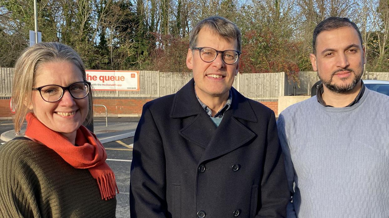 A woman with blonde hair wearing glasses and a green jumper and red scarf smiles towards the camera, she is standing next to a man with grey hair smiling toward the camera. He is wearing glasses and a dark jacket and is standing next to a a bearded man with dark hair who is smiling toward the camera. He is wearing a grey knitted jumper. All three are stood in a line in a car park , wooden fencing and trees can be seen in the background.