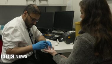 A man wearing a white shirt, tie and blue gloves takes a finger-prick blood sample from a woman seated across a desk, with computer monitors and medical equipment visible in the room.