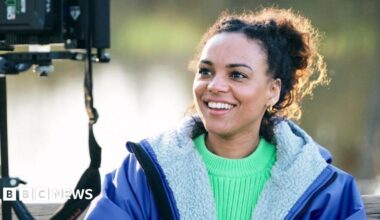 Cat White, a filmmaker, sitting down looking into a camera monitor. She is wearing a bright green jumper with a blue fleece-lined coat over the top. She is smiling but not looking at the camera.