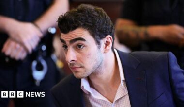 Luigi Mangione, wearing a pink checkered shirt and blue blazer, sits in a New York City courtroom.