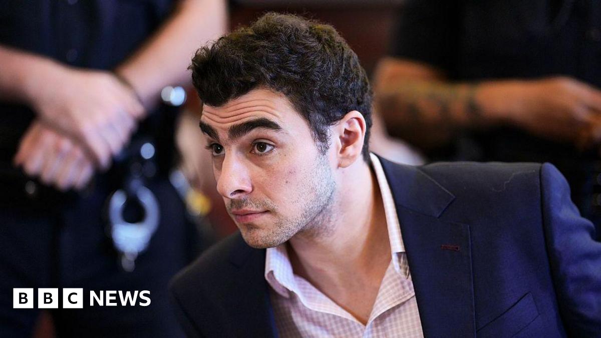 Luigi Mangione, wearing a pink checkered shirt and blue blazer, sits in a New York City courtroom.