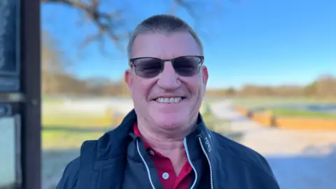GEORGE CARDEN/BBC Alan Covey, a middle-aged man with short hair, wears sunglasses and a blue jacket over a red polo shirt, as he smiles into the camera.