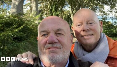 David Letherman and Jim Cunningham pose in a selfie taken by Letherman, who is at the front wearing a black jacket. Cunningham is behind him with his hands on Letherman's shoulders. He is wearing an orange padded jacket and grey scarf. Both men are bald, Letherman has a short grey beard. They are standing in a country lane with hedgerows and trees on each side.