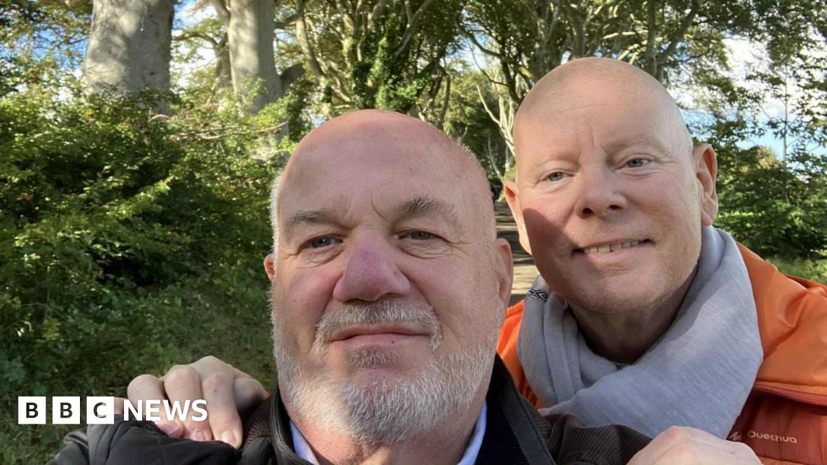 David Letherman and Jim Cunningham pose in a selfie taken by Letherman, who is at the front wearing a black jacket. Cunningham is behind him with his hands on Letherman's shoulders. He is wearing an orange padded jacket and grey scarf. Both men are bald, Letherman has a short grey beard. They are standing in a country lane with hedgerows and trees on each side.