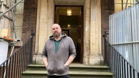 BBC A man with a shaved head and light-brown beard stands in front of the grand stone entrance to a red-brick Georgian house. He is wearing a grey hoody and green polo shirt and has his hands in his pockets. Scaffolding can be seen to either side of the doorway.