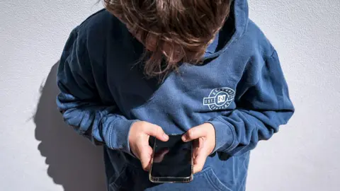 AFP via Getty Images A boy in a blue hoodie looks down at his black phone he is holding in his hands, the screen is blank