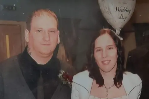 Family handout A man stands to the left of a woman. She is beneath a balloon which says "Wedding Day" and is wearing white. Her red-brown hair falls below her shoulders on to her front, where it touches a chain she is wearing. The man has thinning mousey hair which is cut short. He has a tattoo on his neck. He is wearing a black shirt and a dark grey tie and cardigan